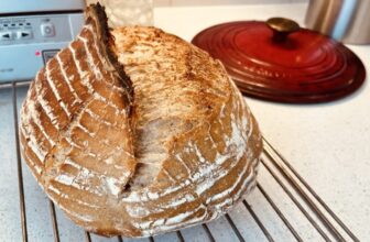 Sourdough loaf on a rack on a kitchen counter, in front of a Dutch oven lid and a square toaster oven.