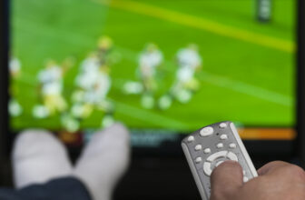 Hand holding a tv remote control. TV has a blurry image of people playing football