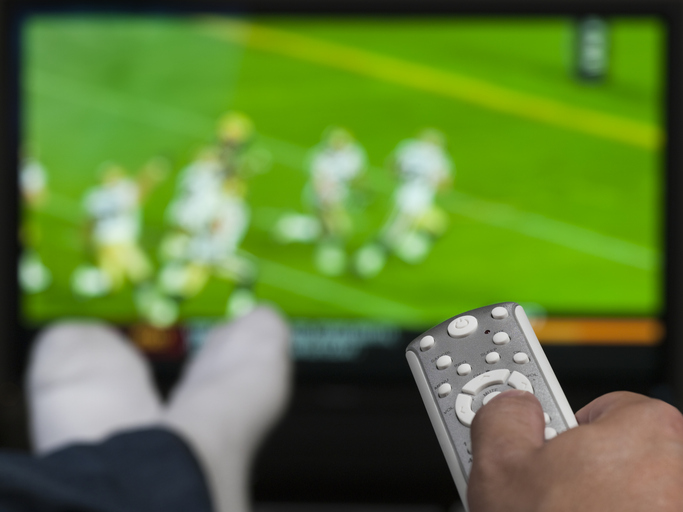 Hand holding a tv remote control. TV has a blurry image of people playing football