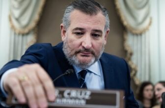 Senator Ted Cruz at a Senate committee hearing, sitting in his seat and using his hand to move a nameplate that says "Mr. Cruz, Chairman."